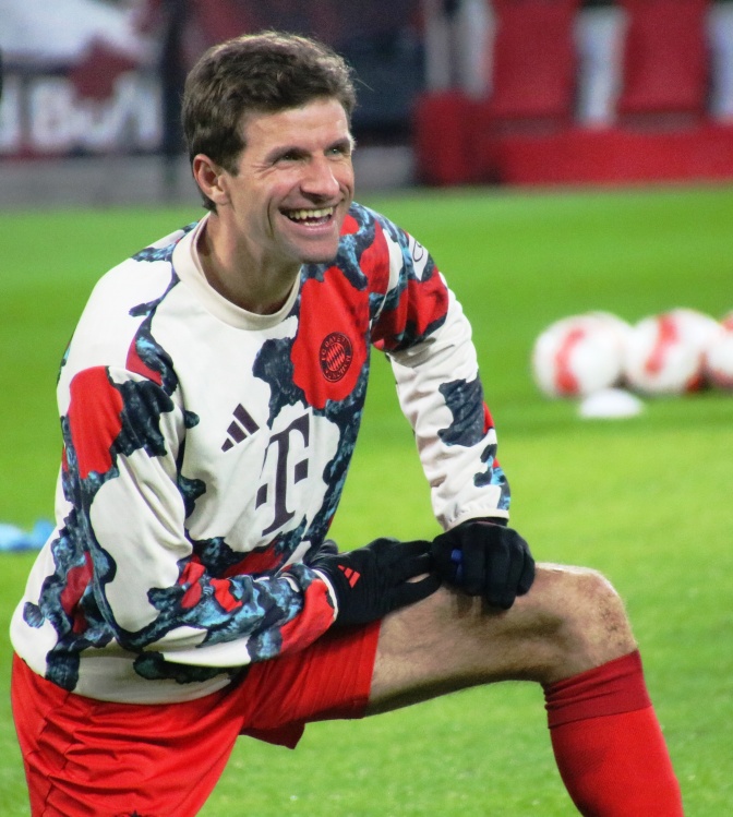Thomas Müller kniet lächelnd im Trikot auf dem Fußballplatz.