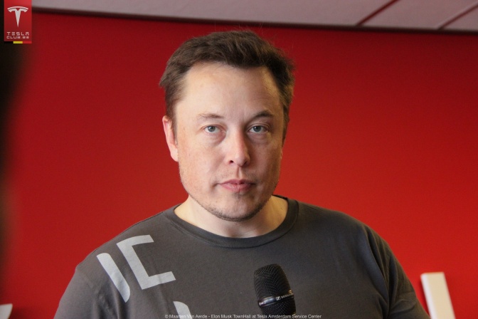 Elon Musk in einem grauen Shirt vor einer roten Wand. Er schaut ernst direkt in die Kamera.