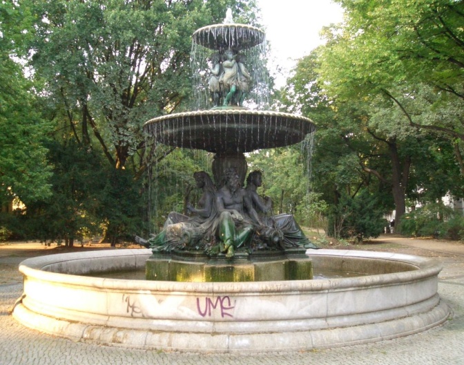 Ein runder Brunnen, außen aus hellem Marmor, innen aus grünlich angelaufenem Kupfer.  In der Mitte sitzen 4 große Figuren aus hellem Stein.