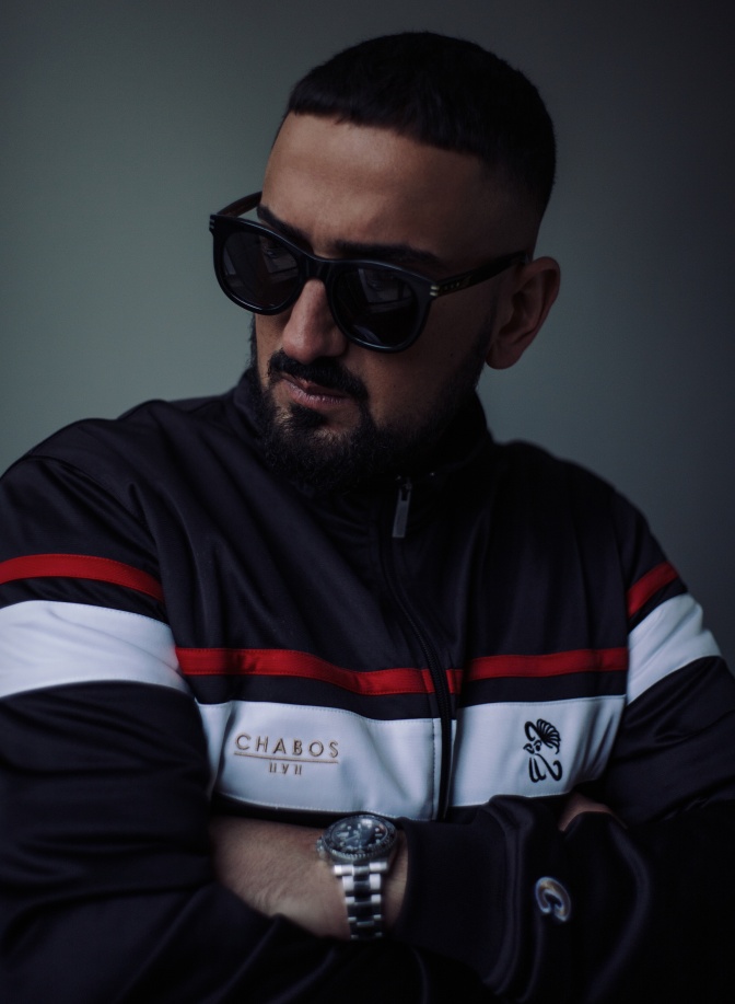 Der Rapper Haftbefehl  mit großer schwarzer Sonnenbrille in einer Sportjacke. Er hat die Arme vor der Brust verschränkt.