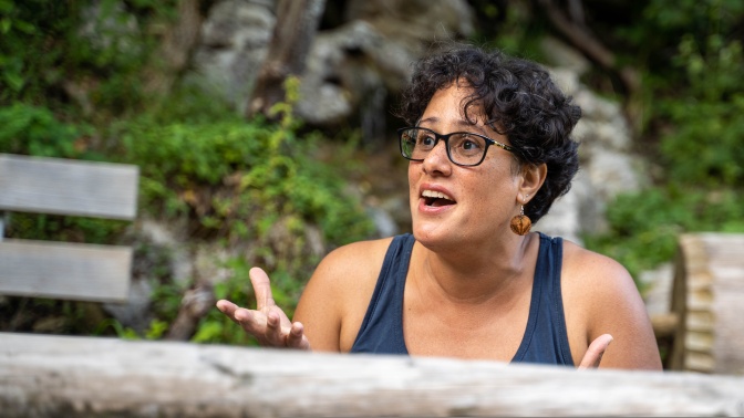 Eine BiPoc Frau mit gewellten, kurzen schwarzen Haaren und schwarzer Brille. Sie schaut sehr überrascht und gestikuliert mit den Händen.