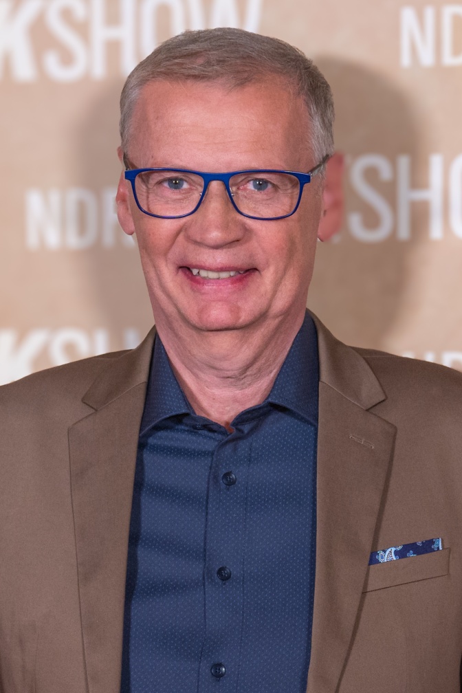 Günther Jauch in einem blauen Hemd und braunem Sakko vor einer Logowand.