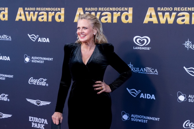 Barbara Schöneberger in einem engen schwarzen Samtkleid vor einer Logowand.