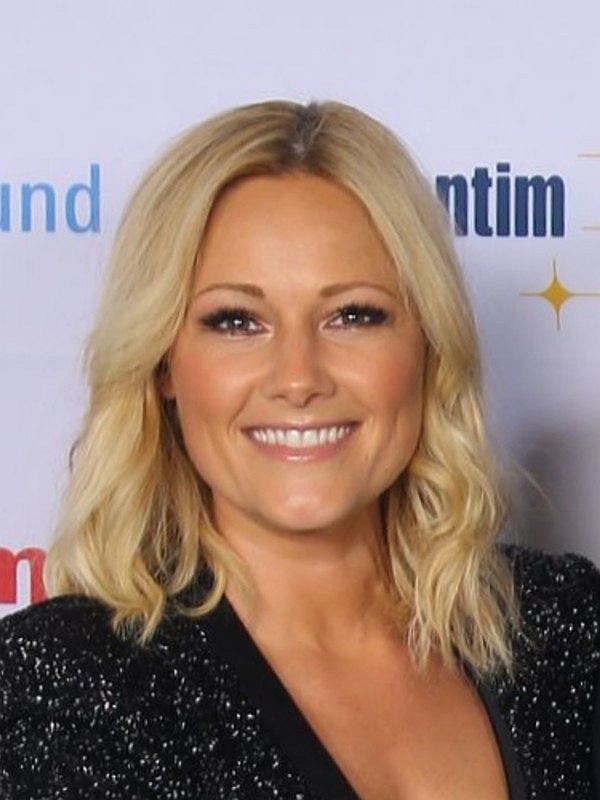 Helene Fischer mit schulterlangen gewellten blonden Haaren. Sie trägt ein schwarzes Glitzerkleid und steht lächelnd vor einer Logowand.
