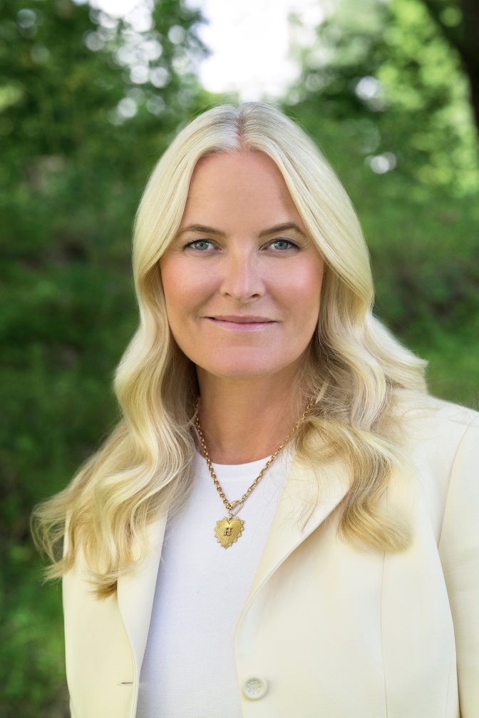Kron·prinzessin Mette-Marit in einem hellgelben Blazer mit langen, gewellten blonden Haaren. Sie schaut direkt in die Kamera und lächelt leicht.