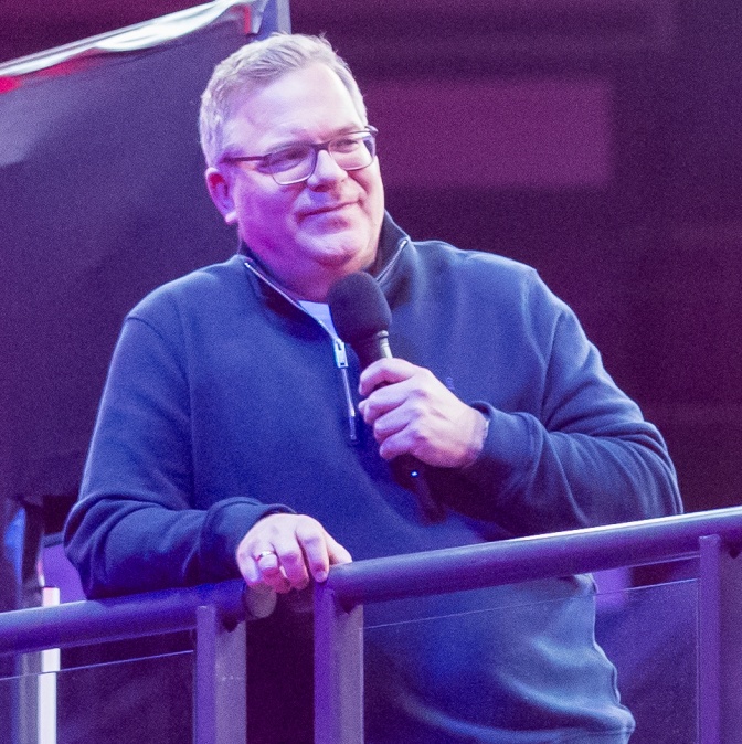 Elton lehnt in einem Wollpulli an einem Geländer und spricht in ein Mikrofon.