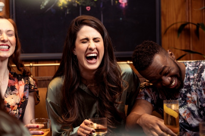 2 weiße Frauen und ein schwarzer Mann lachen ausgelassen. Alle halten einen Drink in der Hand.