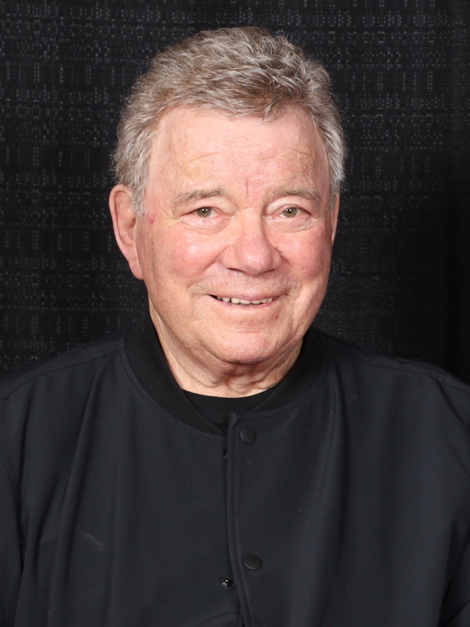 William Shatner in einem schwarzen Shirt vor schwarzem Hintergrund. Er lächelt leicht in die Kamera.