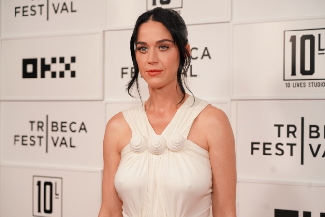 Katy Perry in einem weißen Kleid und mit hochgesteckten dunklen Haaren. Sie steht mit ernstem Blick vor einer Logowand.