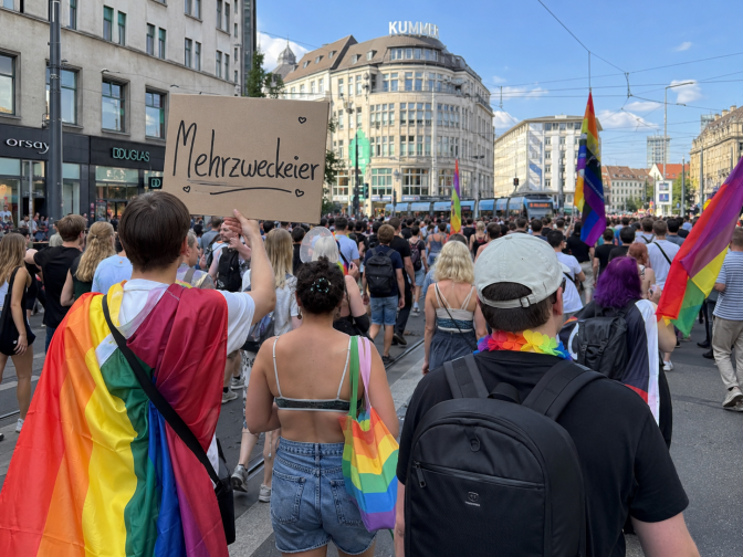 Eine Gruppe von Demonstrierenden bei einem CSD. Sie tragen Regenbogenflaggen. Die Demonstrant*innen sind von hinten zu sehen. Eine Person trägt ein Demoschild mit der Aufschrift Mehrzweckeier.