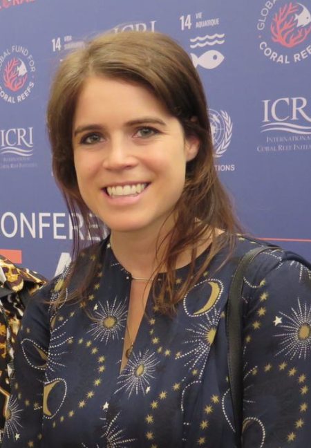 Prinzessin Eugenie hat ihre braunen Haare zurückgebunden und steht lächelnd vor einer Logowand.