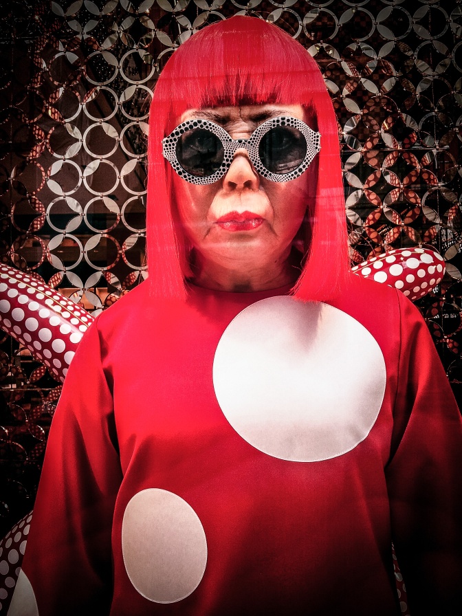Yayoi Kusama mit einer roten Perücke in einem roten Kleid mit weißen Punkten. Die Figur trägt eine schwarze Sonnenbrille. Auch auf der Sonnenbrille und im Hintergrund sind Punkte.