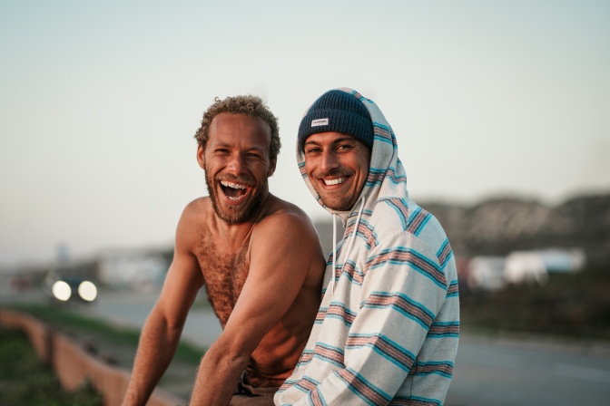 2 lachende Männer am Strand. Einer von beiden trägt ein Hoodie, der andere hat einen nackten, behaarten Oberkörper.
