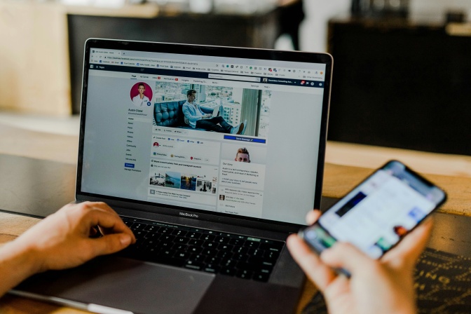 Ein aufgeklappter Laptop, auf dem Facebook geöffnet ist. Außerdem hat die Person ein Handy in der Hand.