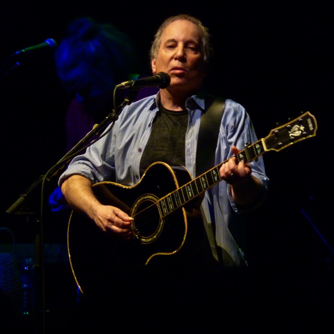 Paul Simon mit grauen Haaren und Stirnglatze. Er spielt Gitarre und singt in ein Mikrofon.