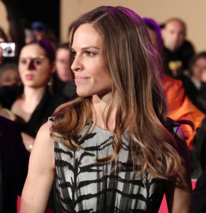 Hilary Swank im Seitenprofil auf dem roten Teppich. Sie trägt ein Abendkleid, im Hintergrund sieht man eine Menschenmenge.