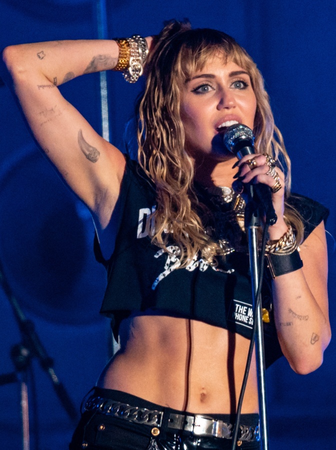 Miley Cyrus in einem crop Top auf der Bühne. Sie hat einen Arm hinter dem Kopf verschränkt und singt in ein Mikro.