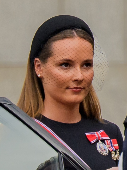 Ingrid Alexandra trägt einen schwarzen Haarreif mit einem kleinen Schleier und einen Orden. Sie steht aufrecht und mit ernstem Blick neben einem Auto.