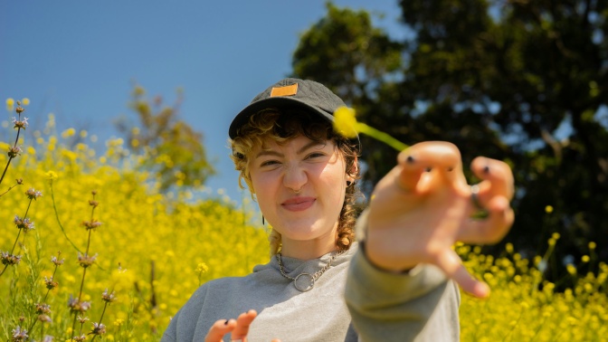Eine Frau mit Schirmmütze steht vor einem Rapsfeld. Sie hat eine Rapsblüte gepflückt und streckt sie in die Kamera.