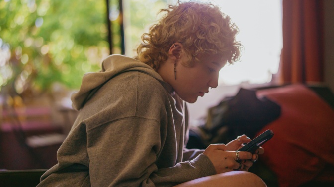 Eine Frau mit hellblonden kurzen Locken sitzt über ihr Handy gebeugt. Sie ist im Seitenprofil zu sehen.