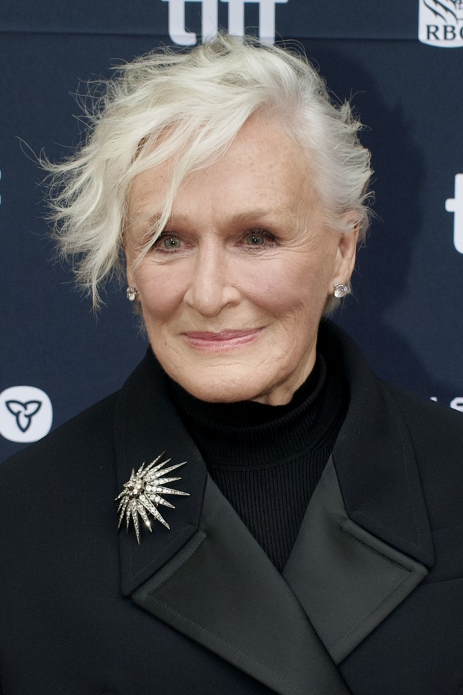Glenn Close mit weißen Haaren und einer asymmetrischen Frisur. Sie steht in schwarzer Kleidung vor einer Logowand.
