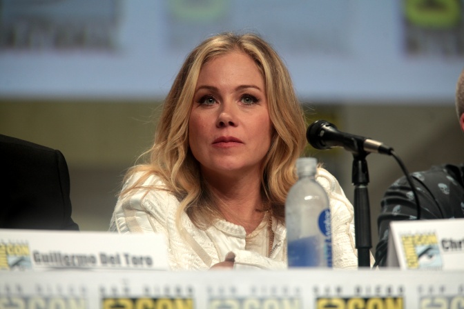 Christina Applegate sitzt vor einer Reihe Namensschilder an einem Mikrofon. Sie trägt ein weißes Oberteil und hat gewellte, blonde Haare.