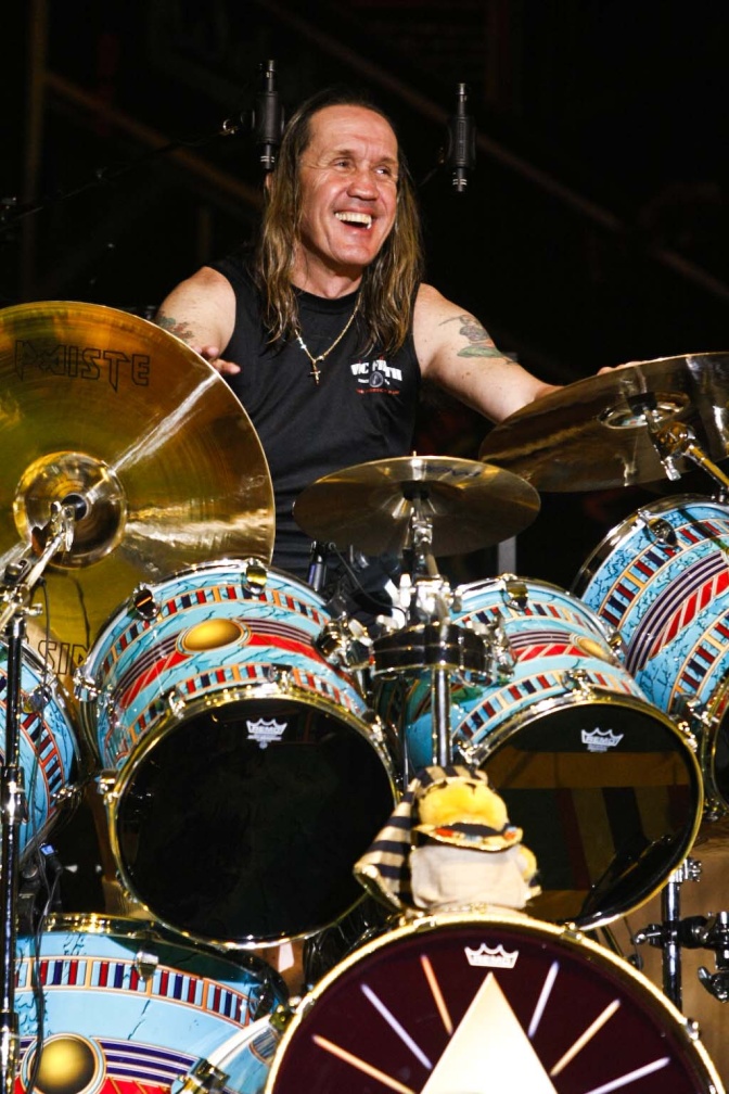 Nicko McBrain mit langen Haaren in einem schwarzen Tank Top. Er lächelt und sitzt hinter seinem Schlagzeug.
