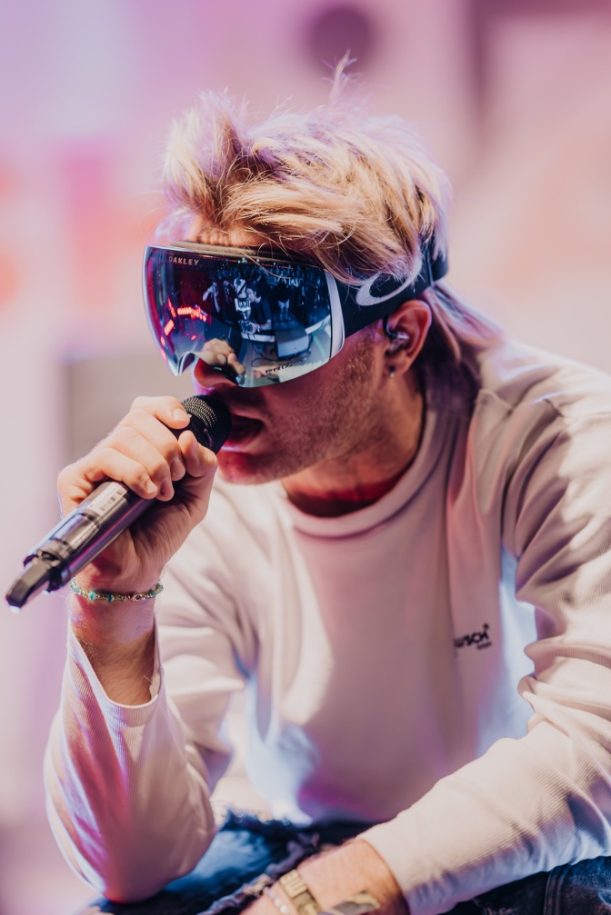 Ski Aggu  it blondierten Haaren und spiegelnder Skibrille. Er hockt und singt in ein Mikrofon.