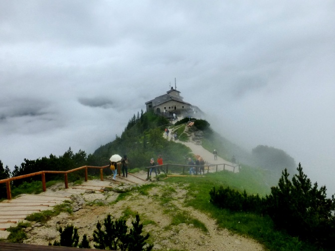 Ein Beggasthof, fast komplett von dichtem Nebel umgeben. Einige Menschen laufen im Vordergrund vorbei.