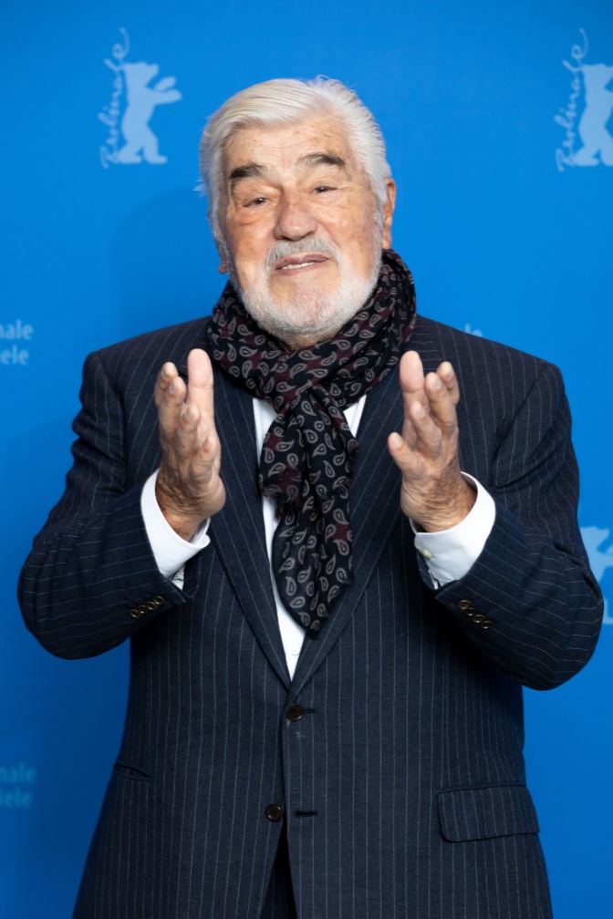 Mario Adorf mit weißen Haaren und weißem Bart. Er steht vor einer Logowand und gestikuliert beim Sprechen mit den Händen.