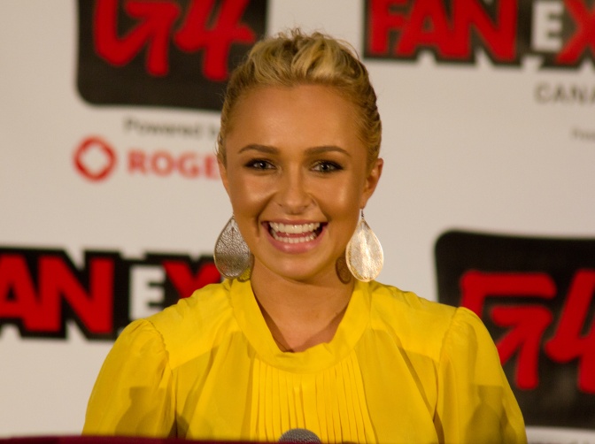 Hayden Panettiere in einer gelben Bluse vor einer Logowand mit dem Schriftzug Expo. Sie sitzt vor einem Mikrofon und lächelt in die Kamera.