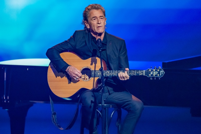 Peter Maffay spielt eine akustische Gitarre. Die Bühne hinter ihm ist blau beleuchtet.