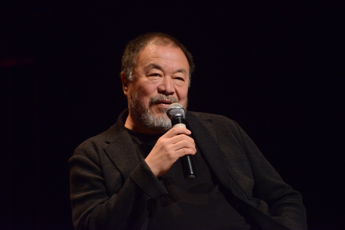Ai Weiwei in schwarzer Kleidung und vor schwarzem Hintergrund. Er spricht in ein Mikrofon.