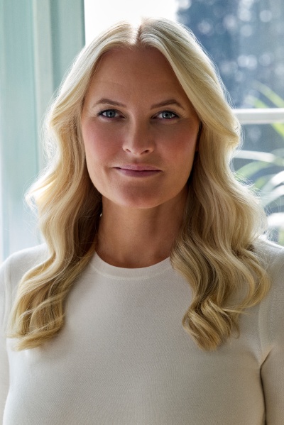 Kronprinzessin Mette Marit mit offenen, gewellten Haaren in einem schlichten, weißen Shirt. Sie steht vor einem Fenster und schaut direkt in die Kamera.