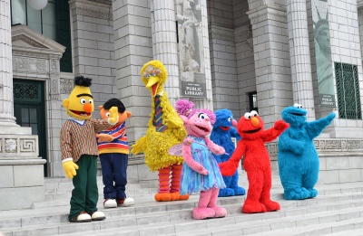 Eine Puppe in einem Bertkostüm steht mit anderen Sesamstraßenfiguren zusammen vor einem Gebäude