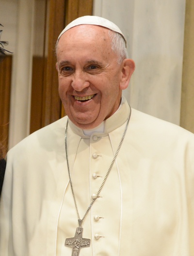 Papst Franziskus lächelt. Er trägt einen weißen Talar und eine goldene Kette mit einem Kreuz um den Hals.