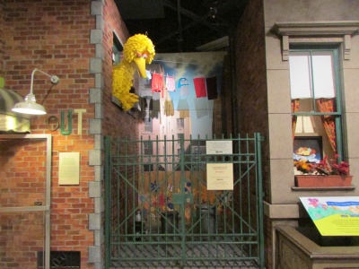 Bibo, ein gelber Vogel, streckt seinen Kopf aus dem Fenster eines Backsteinhauses in einer Fernsehkulisse.