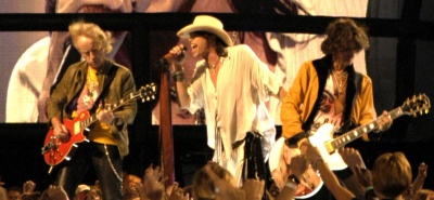 Mitglieder der Band Aerosmith auf der Bühne: Sänger Steven Tyler, Gitarrist Jimmy Crespo und Bassist Tom Hamilton stehen zusammen auf der Bühne, Steven Tyler singt, die beiden anderen spielen ihre Instrumente. Im Vordergrund sieht man die Hände jubelnder Fans, im Hintergrund eine Videoleinwand, auf die das Konzert projeziert wird.