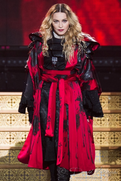 Madonna in einem rot-schwarzen Kleid auf der Bühne. Sie hat lange, gewellte, blonde Haare, die sie offen trägt.  Sie geht eine Treppe herunter.