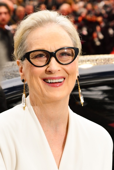Meryl Streep in einer hellen Bluse mit auffälliger schwarzer Brille. Sie lacht und schaut direkt in die Kamera.