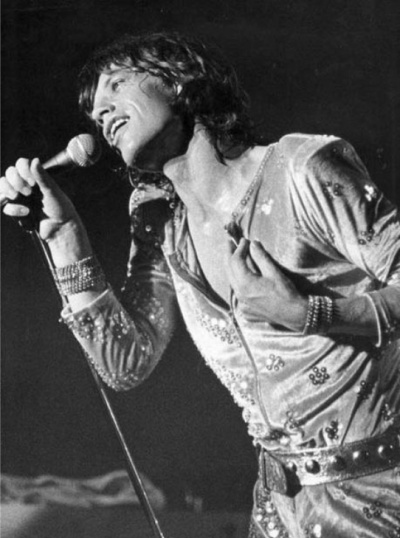 Ein schwarz-weiß-Foto von Mick Jagger auf der Bühne. Er singt in ein Mikrophon und greift sich mit einer Hand ans Herz.