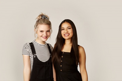 2 junge Frauen stehen lächelnd nebeneinander. Eine von beiden hat blonde Haare zum Dutt frisiert. Die andere der beiden hat glatte, lange, dunkle Haare.