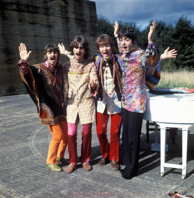 Die 4 Mitglieder der Beatles heben die Arme in die Luft und jubeln. Sie stehen vor einem weißen Klavier.