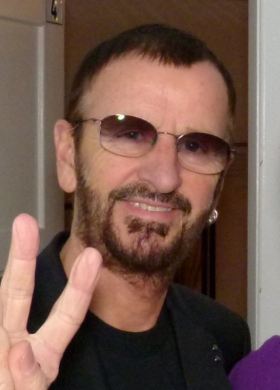 Ringo Starr mit Sonnenbrille. Er macht das Peace-Zeichen mit den Fingern und lächelt.