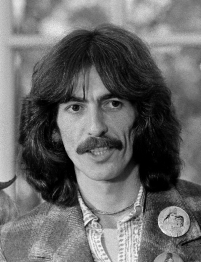 George Harrison mit kinnlangen Haaren und Schnäuzer. Er trägt ein kariertes Sakko und einen Button.