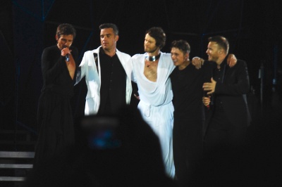 Die 5 Bandmitglieder stehen nebeneinander und singen in Mikrophone.