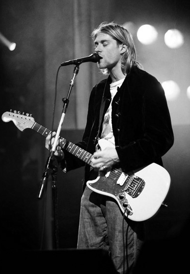 Kurt Cobain auf der Bühne. Er spielt eine weiße E-Gitarre und singt mit geschlossenen Augen in ein Mikrophon.