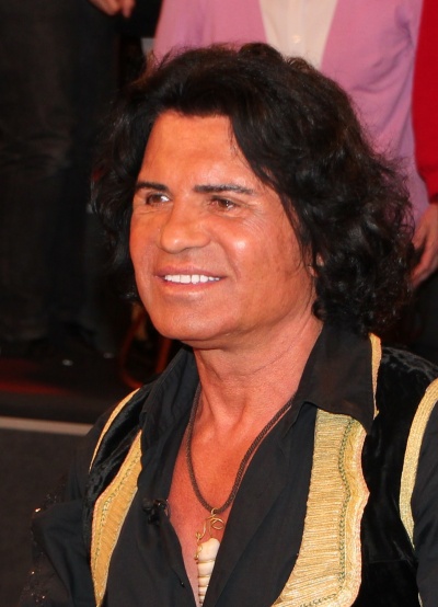 Costa Cordalis  mit schwarzem Hemd. Er hat halblange, gewellte Haare und lächelt.