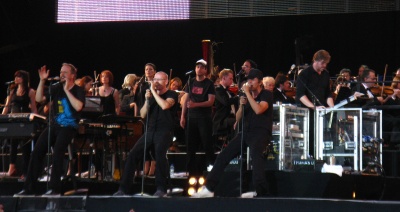 Die Fantastischen Vier in schwarzer Kleidung auf der Bühne. Sie stehen vor Standmikrophonen. Hinter ihnen sieht man ein klassisches Orchester,