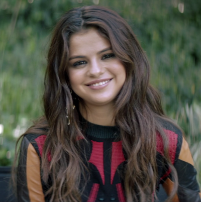 Selena Gomez mit langen braunen Haaren und Mittelscheitel. Sie trägt einen Strickpullover.